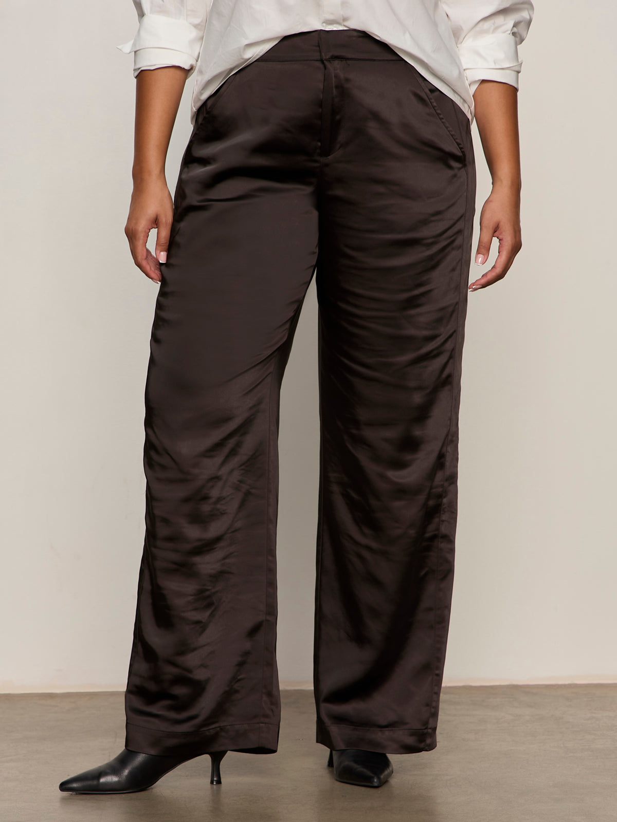 A person wearing Sanctuary Clothing's modern satin trouser in black (extended sizing), paired with a white button-up shirt and black pointed-toe heels, stands against a plain background.