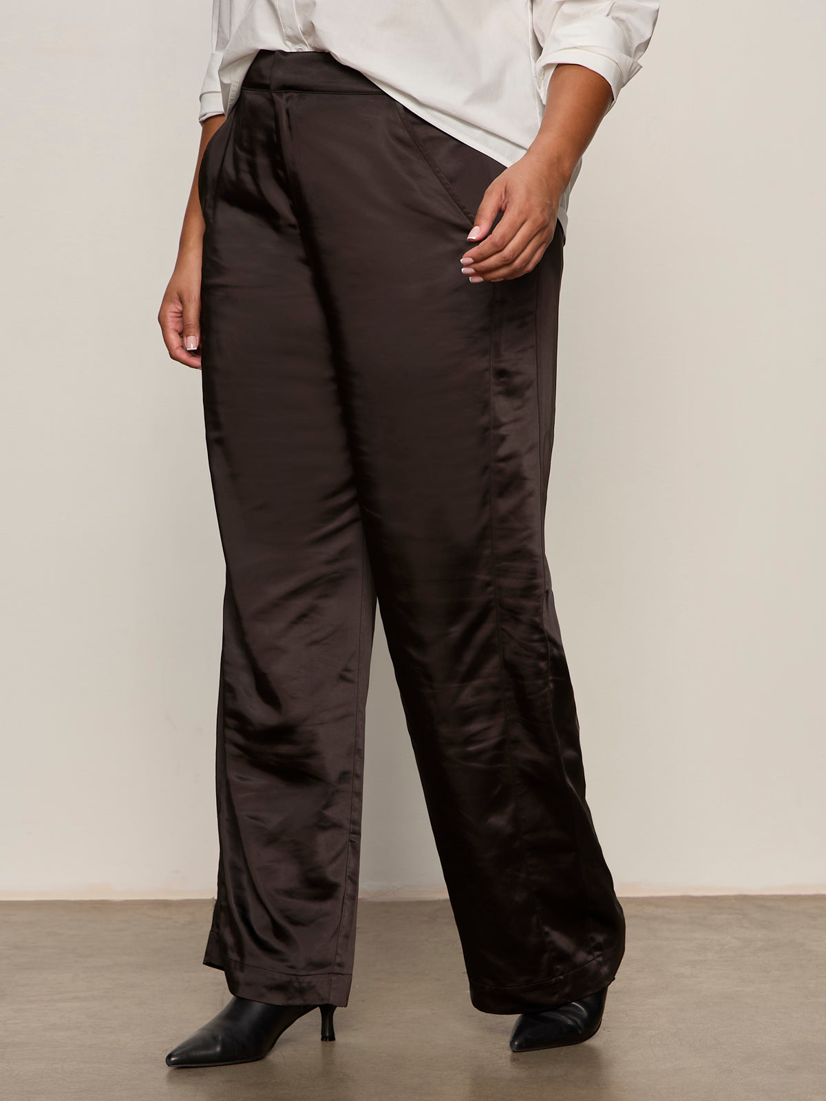 A person stands against a plain background, shown from the shoulders down, wearing Sanctuary Clothing's modern satin trouser in black extended sizing, paired with a white rolled-sleeve shirt and black pointed-toe heeled boots.