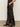 A person models the modern satin trouser black from Sanctuary Clothing, styled with a black top and pointed shoes, standing sideways with one hand in the pocket against a plain background.