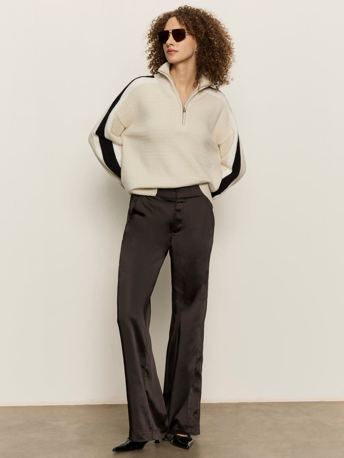 Wearing sunglasses, a cream zip-neck sweater with black and white sleeve stripes, Sanctuary Clothing's modern satin trouser in black, and pointed black shoes, a curly-haired person stands before an off-white wall.