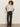 Wearing sunglasses, a cream zip-neck sweater with black and white sleeve stripes, Sanctuary Clothing's modern satin trouser in black, and pointed black shoes, a curly-haired person stands before an off-white wall.
