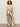 A woman with curly hair wears the Sanctuary Clothing cadence relaxed straight pant in platinum, paired with a sheer lace top, belt, and beige heeled boots, standing against a plain background.