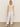 Someone with long hair is wearing a sleeveless beige knit top, paired with Sanctuary Clothing's "relaxed linen cruiser semi high rise pant" in bleached white and beige shoes, against a plain light-colored background.