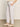 A person wearing Sanctuary Clothing's relaxed linen cruiser semi high rise pant in bleached white stands with one hand in a pocket. The light beige crocheted flats and the simple, neutral-colored wall and floor complete the scene.