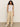 A person with long curly hair stands against a neutral background, wearing a white sleeveless top, beige Sanctuary Clothing's relaxed linen cruiser semi high rise pants in sandstone, and white sneakers. Their hands are in their pockets as they smile slightly at the camera.