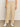 A person is wearing Sanctuary Clothing's relaxed linen cruiser semi high rise pant in sandstone with large front pockets, white sneakers, and a tucked-in white shirt. The background features an off-white wall and gray floor. The pants have a relaxed, slightly cropped fit.