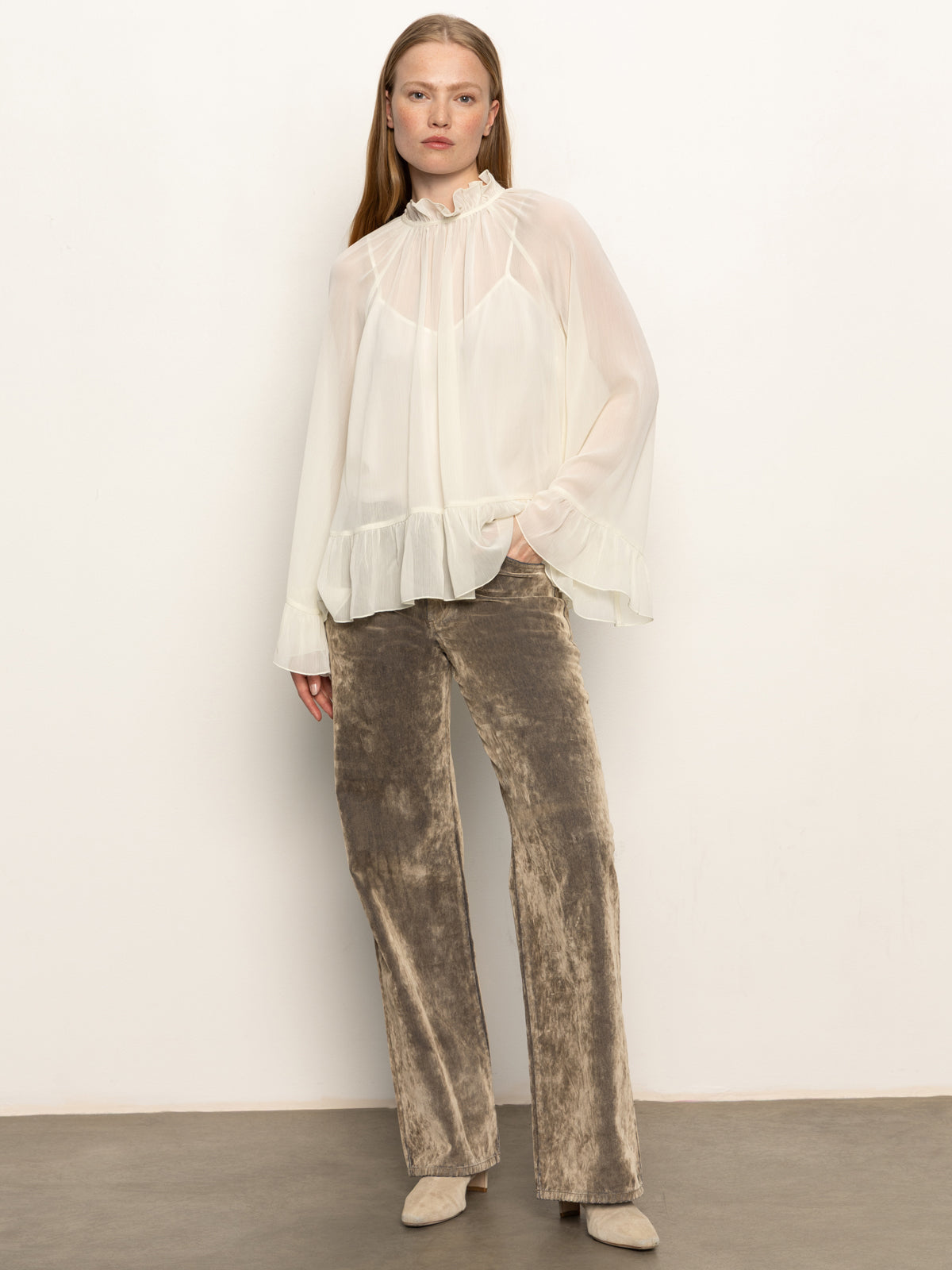 A woman poses against a plain wall wearing Sanctuary Clothing’s cadence relaxed straight jean in light bronze, paired with a sheer, long-sleeve white ruffled blouse and beige shoes, one hand casually placed in her pocket.