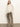 A woman poses against a plain wall wearing Sanctuary Clothing’s cadence relaxed straight jean in light bronze, paired with a sheer, long-sleeve white ruffled blouse and beige shoes, one hand casually placed in her pocket.