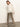 A woman stands against a plain wall, wearing a sheer white ruffled blouse and the Sanctuary Clothing cadence relaxed straight jean in light bronze, paired with matching heeled boots. She looks down with one arm by her side.