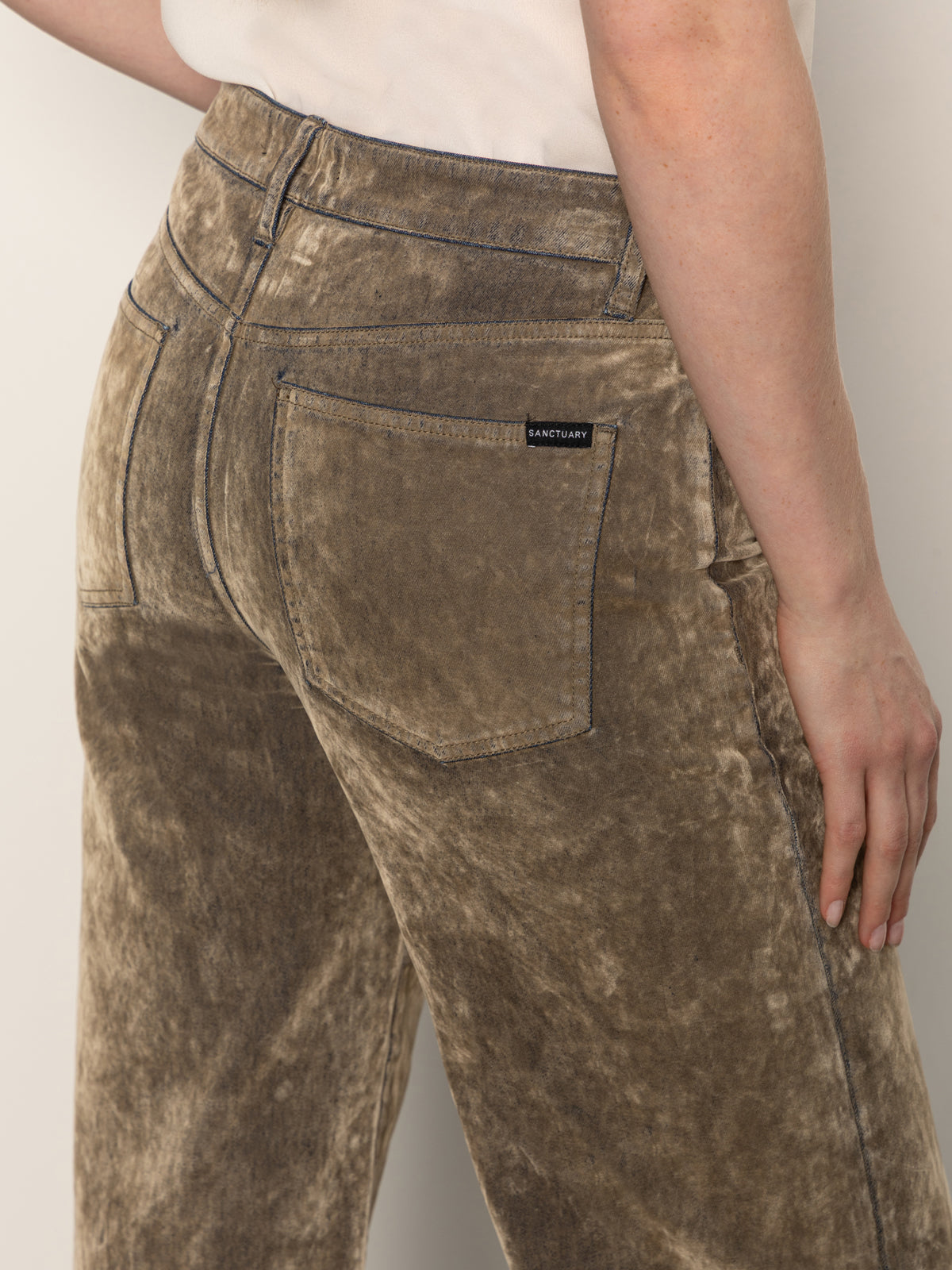 A person wearing Sanctuary Clothing's cadence relaxed straight jean in light bronze, featuring a high waist, visible back pockets, and a small "Sanctuary" label on the right pocket, stands with their right hand at their side.