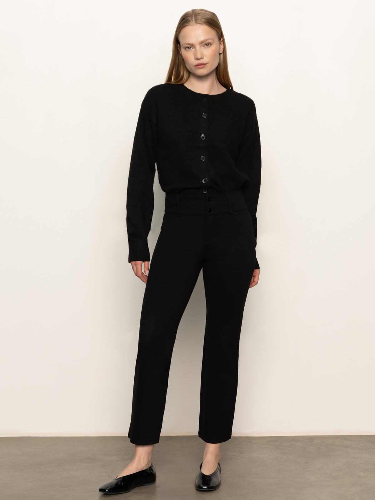 A woman with straight, light brown hair and a neutral expression stands against a plain wall, wearing Sanctuary Clothing's 90s straight legging black pants with a black button-up cardigan and black flats.