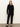 A woman with straight, light brown hair and a neutral expression stands against a plain wall, wearing Sanctuary Clothing's 90s straight legging black pants with a black button-up cardigan and black flats.