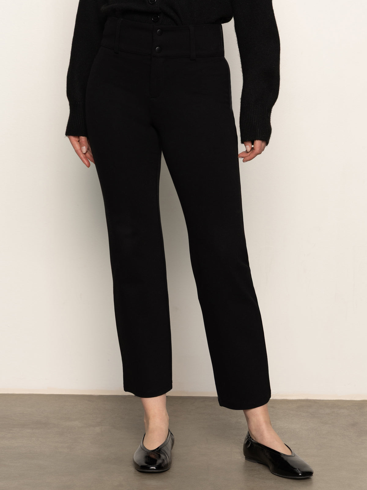 A person stands against a white wall wearing Sanctuary Clothing's 90s straight legging black pants, a black long-sleeve sweater, and black flats. Their face is not visible in the image.