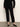A person stands against a white wall wearing Sanctuary Clothing's 90s straight legging black pants, a black long-sleeve sweater, and black flats. Their face is not visible in the image.