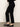 A person models the Sanctuary Clothing 90s Straight Legging Black pants, paired with shiny black flats, while standing against a plain light-colored wall; their upper body is out of frame.