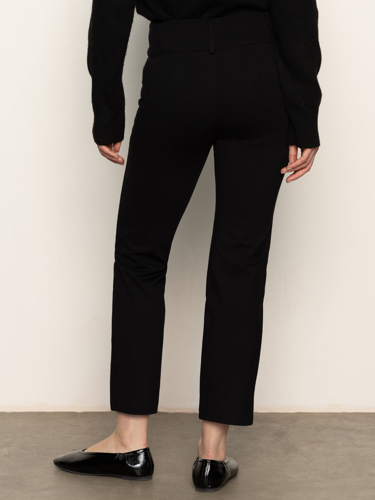 Someone is standing with their back to the camera against a light wall, wearing Sanctuary Clothing’s 90s straight legging black pants, paired with a black long-sleeve top and black shiny flats.
