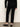 Someone is standing with their back to the camera against a light wall, wearing Sanctuary Clothing’s 90s straight legging black pants, paired with a black long-sleeve top and black shiny flats.
