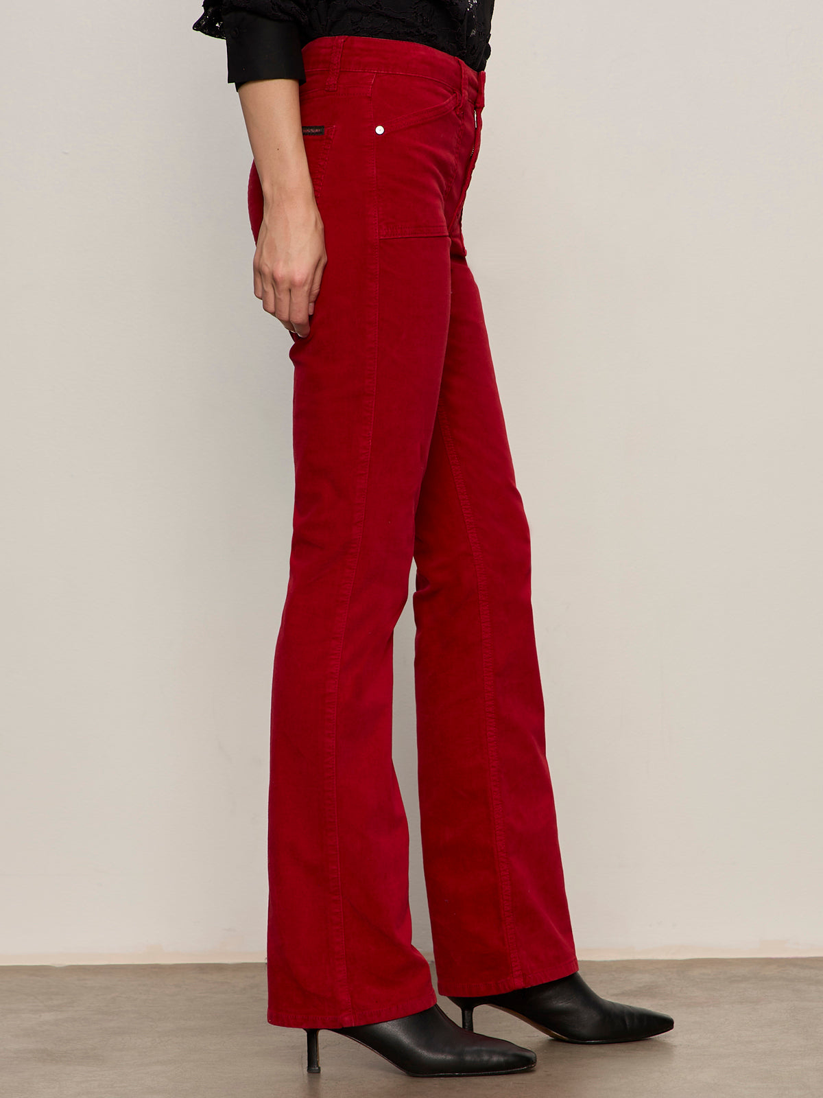 A person stands sideways against a light wall, wearing Sanctuary Clothing's velveteen brook bootcut pant in cherry lacquer and black heeled boots, with their left arm relaxed by their side.