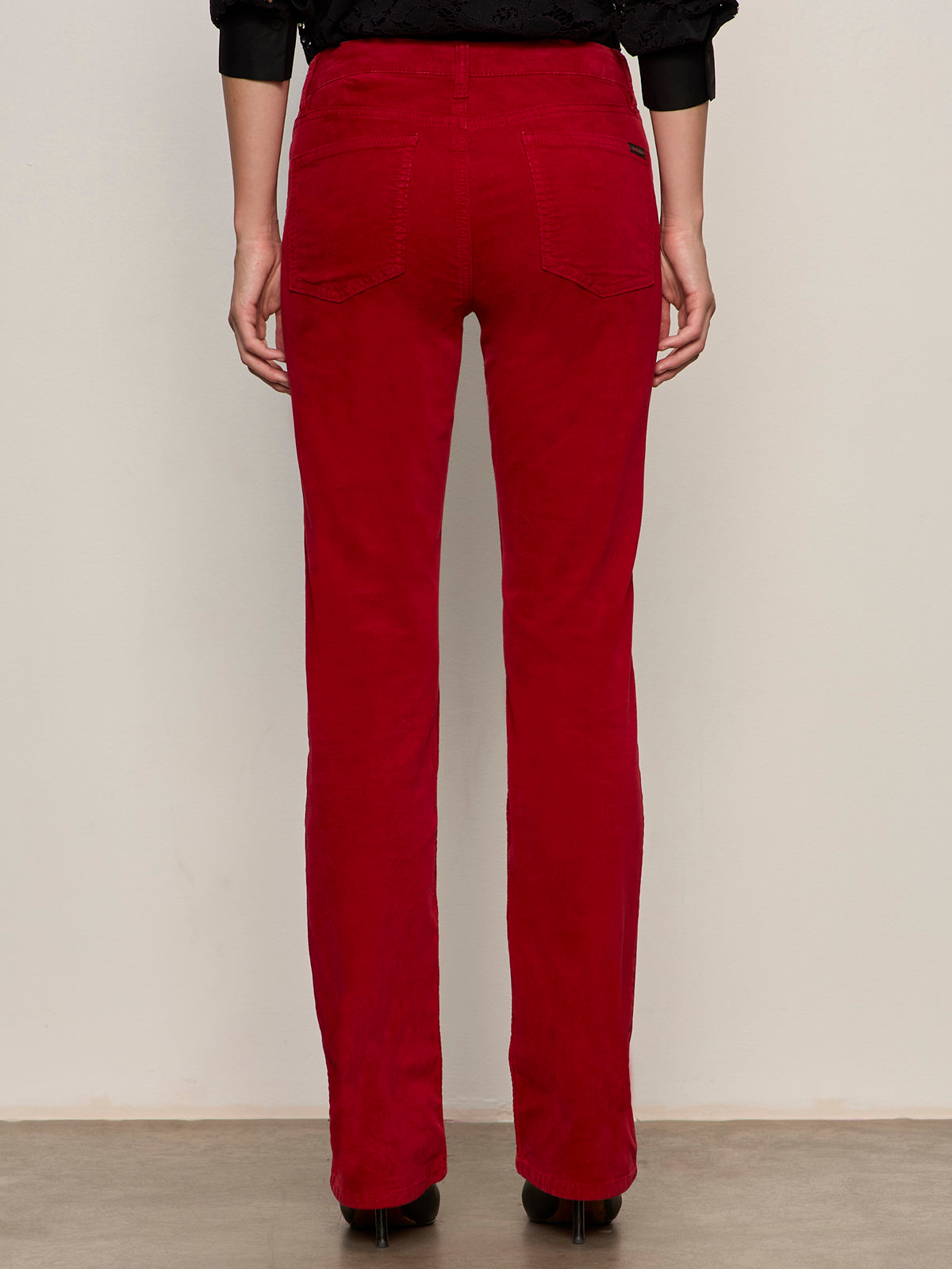 A person stands with their back to the camera, wearing Sanctuary Clothing„¢s velveteen brook bootcut pant in cherry lacquer and black high-heeled shoes, set against a plain light-colored wall.