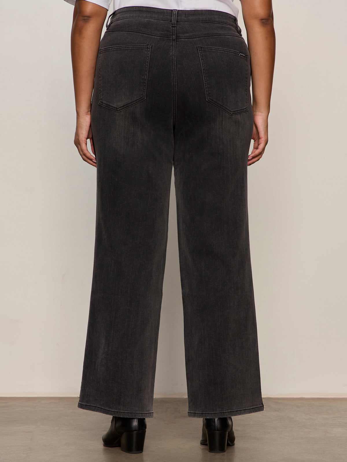 A person stands facing away from the camera wearing Sanctuary Clothing's tuxedo relaxed straight jean black tie extended sizing and black shoes against a neutral background.