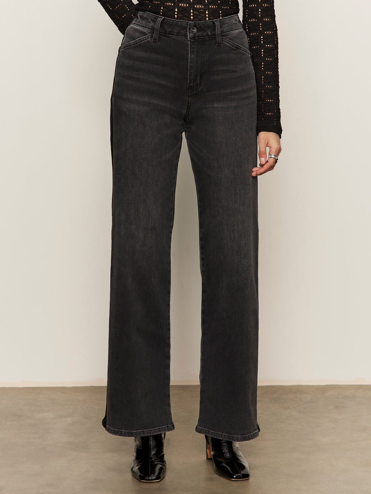A person wearing Sanctuary Clothing's tuxedo relaxed straight jean black tie with a textured black long-sleeve top and heeled boots, standing against a plain light background.