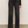 A person wearing Sanctuary Clothing's tuxedo relaxed straight jean black tie with a textured black long-sleeve top and heeled boots, standing against a plain light background.