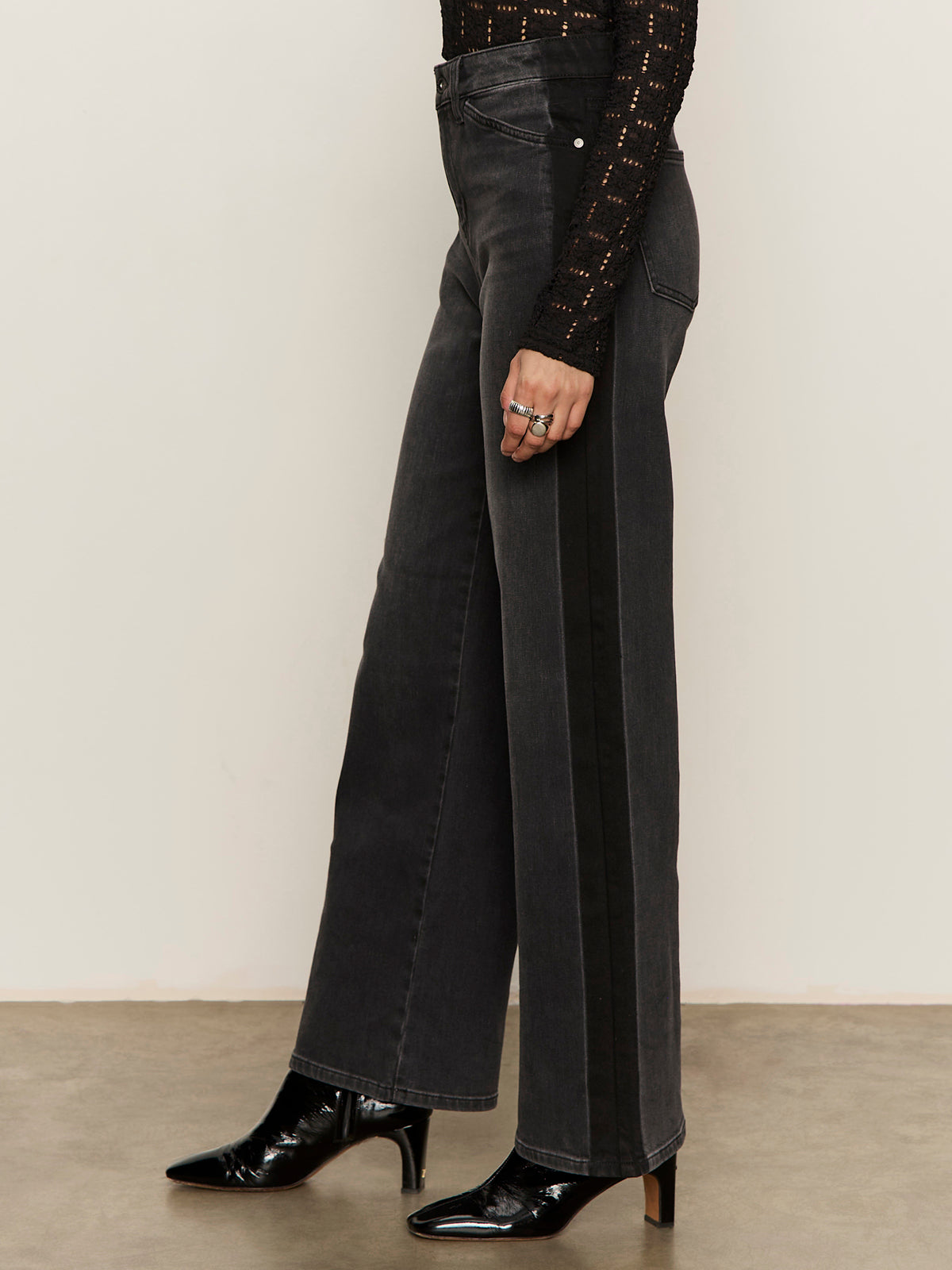 A person wearing Sanctuary Clothing„¢s tuxedo relaxed straight jean black tie, a black textured long-sleeve top, black pointed-toe heeled boots, and silver rings stands on a concrete floor.