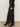 A person wearing Sanctuary Clothing„¢s tuxedo relaxed straight jean black tie, a black textured long-sleeve top, black pointed-toe heeled boots, and silver rings stands on a concrete floor.
