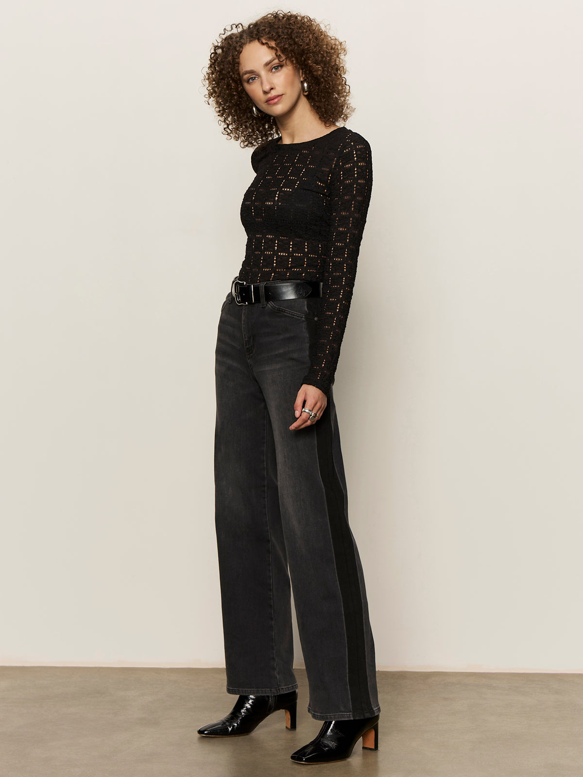 A woman with curly hair wears the Sanctuary Clothing tuxedo relaxed straight jean black tie, a black long-sleeve sheer knit top, black belt, and black heeled boots, standing against a plain light background.