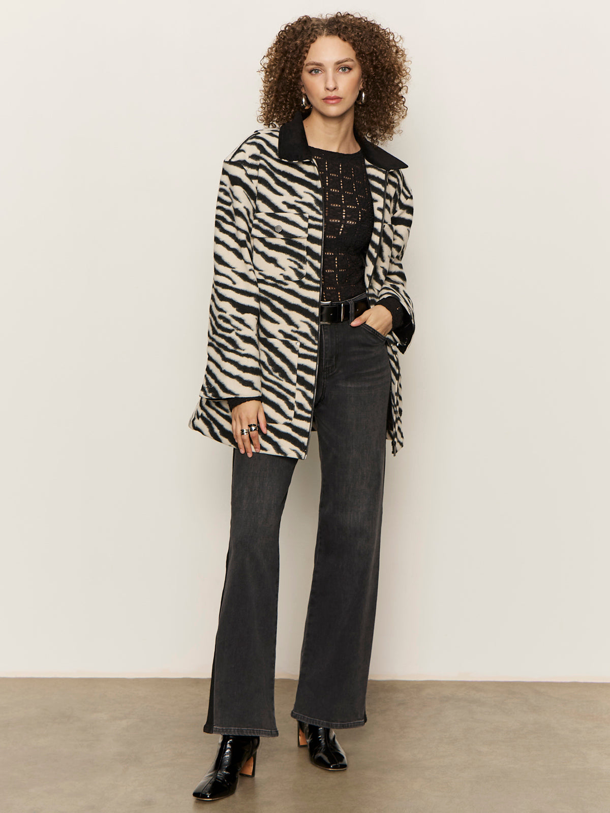 A woman with curly hair wears Sanctuary Clothing„¢s tuxedo relaxed straight jean black tie, a black knit top, zebra print coat, black belt, and heeled boots, standing against a plain light-colored wall.