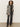 A woman with curly hair wears Sanctuary Clothing„¢s tuxedo relaxed straight jean black tie, a black knit top, zebra print coat, black belt, and heeled boots, standing against a plain light-colored wall.