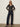 A woman wearing black sunglasses, a black floral long-sleeve shirt, and Sanctuary Clothing's marine full length jean first sight stands against a beige wall with her hands in her pockets.
