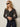 A woman with long blonde hair and black sunglasses stands in a relaxed pose against a light wall, wearing Sanctuary Clothing's marine full length jean first sight, paired with a black floral button-up blouse and black belt.