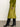 A woman stands indoors on a concrete floor against an off-white wall, wearing Sanctuary Clothingžs everyday midi skirt in martini olive, paired with a green sweater and black knee-high heeled boots.