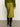 A person stands on a concrete floor facing away, dressed in a green sweater, black knee-high boots, and the Sanctuary Clothing everyday midi skirt in martini olive.