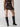 Rear view of a person showcasing the Sanctuary Clothing "Night Out Sequin Skirt Night Spots," a shiny, leopard-print mini skirt, paired with black high-heeled knee-high boots. The photo captures the legs and lower torso against a plain, light gray background.