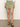 A person is wearing a "all buttoned up skirt sage" by Sanctuary Clothing, featuring large pockets. Their feet are in strappy brown sandals. Only the lower half of the body is visible against a plain, light-colored backdrop on a concrete floor.