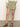 A person in a white textured blouse and the all buttoned up skirt in sage from Sanctuary Clothing stands against a neutral background, with hands in pockets and wearing brown sandals.