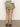 A rear view of someone in Sanctuary Clothing's "all buttoned up skirt" in sage, paired with a textured white long-sleeve blouse and brown flat sandals. The setting is a neutral indoor space with a concrete floor.