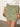 A person wearing a white embroidered blouse and Sanctuary Clothing's 'all buttoned up skirt sage' has their hands resting by their sides, adorned with a simple bracelet. The skirt features large front pockets, and the background is neutral.