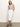 A person with long hair poses against a plain background, wearing a striped sleeveless top and slipping their hands into the pockets of their carrier denim skirt in bleached white by Sanctuary Clothing. They complete the look with black sandals.