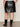 Someone wearing the Sanctuary Clothing camden skirt black extended sizing and black knee-high boots stands with their back to the camera, facing a neutral wall on a muted gray floor.