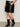 Someone wearing the Sanctuary Clothing camden skirt black extended sizing, a white top, and black mid-calf boots stands with one hand in their pocket against a light background.