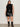 A woman with long brown hair and a confident look stands against a plain background wearing Sanctuary Clothing’s camden skirt black extended sizing, a sleeveless black knit top, and black mid-calf boots.
