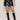 A person models the camden skirt black by Sanctuary Clothing with a black top and knee-high black boots, standing against a plain light background with hands at their sides.