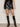 A person models the camden skirt black by Sanctuary Clothing with a black top and knee-high black boots, standing against a plain light background with hands at their sides.