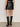 A person stands facing away from the camera, wearing the camden skirt black by Sanctuary Clothing and knee-high black heeled boots against a plain, neutral background.