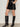 A person models the Sanctuary Clothing camden skirt black, paired with black knee-high heeled boots, against a plain light background. Only the lower half of their body is visible, with arms relaxed at their sides.