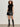 A woman with curly hair wears a sleeveless black turtleneck top, knee-high black boots, and the Camden Skirt Black by Sanctuary Clothing. She stands against a light-colored wall with one hand in her pocket.
