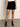 A person wears a white top, black socks, black heeled shoes, and the quilted velvet mini skirt in black by Sanctuary Clothing while standing indoors on a concrete floor against a plain light-colored wall.